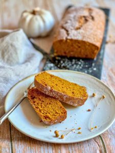 Veganer Kürbiskuchen auf einer schwarzen Platte und zwei Scheiben veganer Kürbiskuchen auf einem Teller.