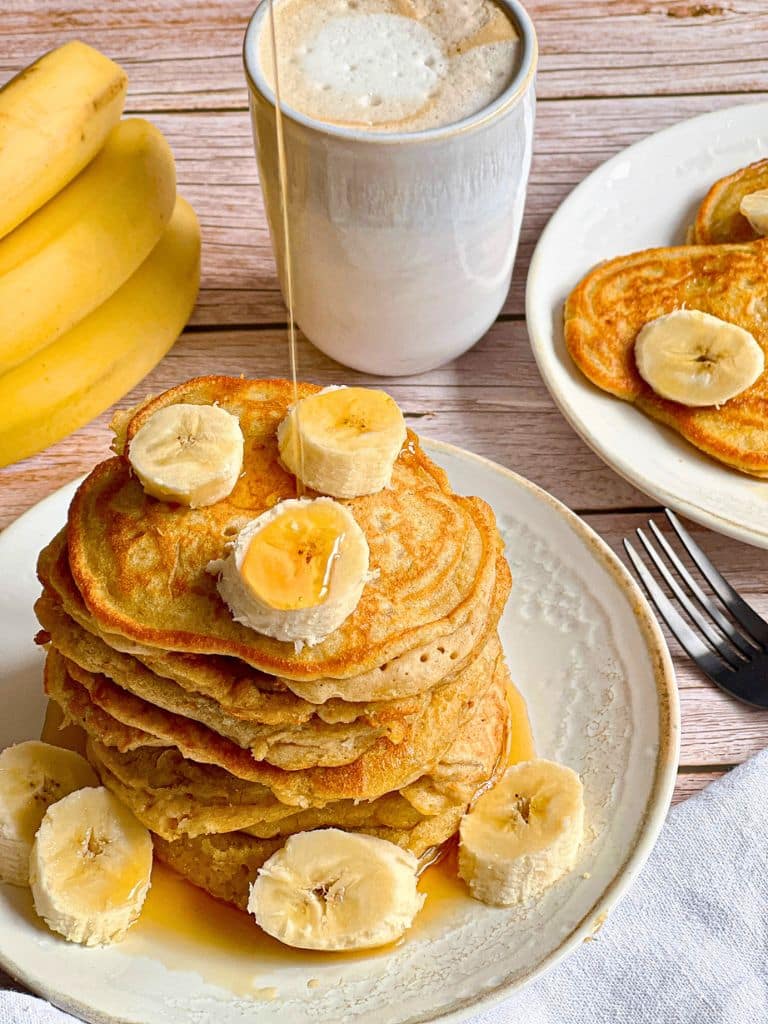 Schnelle vegane Bananen-Pancakes mit Ahornsirup, der von oben übergegossen wird, gestapelt auf einem Teller.