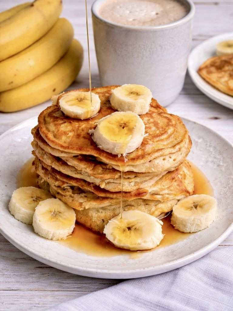 Vegane Bananen-Pancakes mit Ahornsirup, der von oben übergegossen wird, gestapelt auf einem Teller.