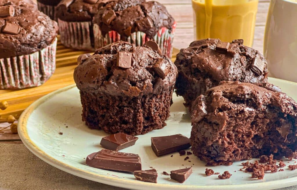 Vegane Schokomuffins (Double Chocolate Muffins) auf einem Teller mit Mandelmus und auf einem Holzbrett, daneben eine Tasse Kaffee.