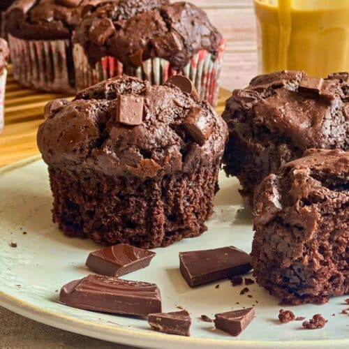 Vegane Schokomuffins (Double Chocolate Muffins) auf einem Teller mit Mandelmus und auf einem Holzbrett, daneben eine Tasse Kaffee.