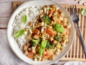 Gemüse mit Tofu in Erdnusss-Soße mit Reis auf einem Teller.