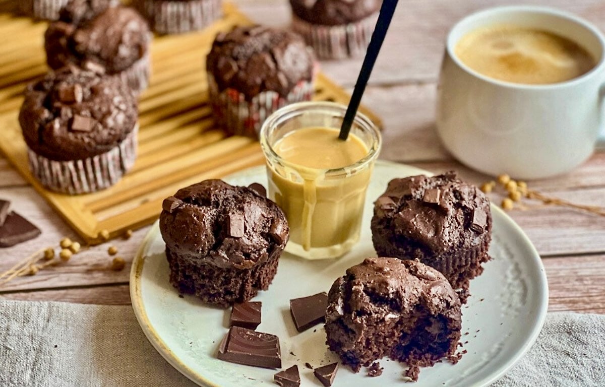 Vegane Schokomuffins (Double Chocolate Muffins) auf einem Teller mit Mandelmus und auf einem Holzbrett, daneben eine Tasse Kaffee.