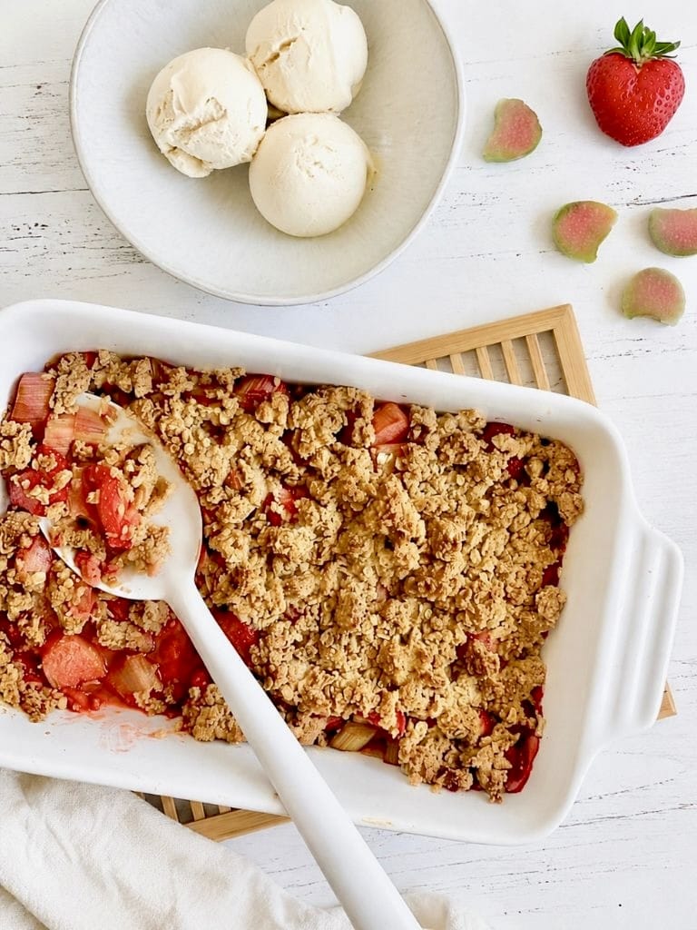 Veganer Rhabarber-Erdbeer-Crumble in einer Auflaufform auf einem Holztisch mit einer Schüssel mit 3 Kugeln Vanilleeis.