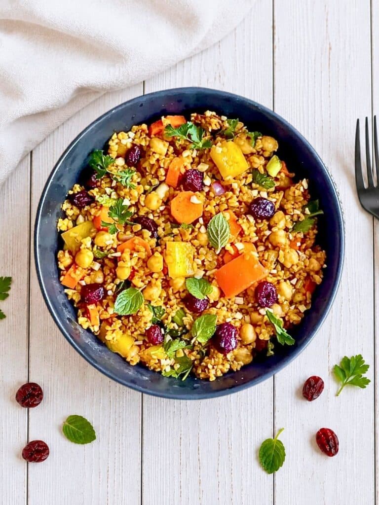 Veganer Bulgursalat mit Gemüse und Kräutern in einer Schüssel auf einem Holztisch, eine Gabel und ein Tuch liegen daneben.