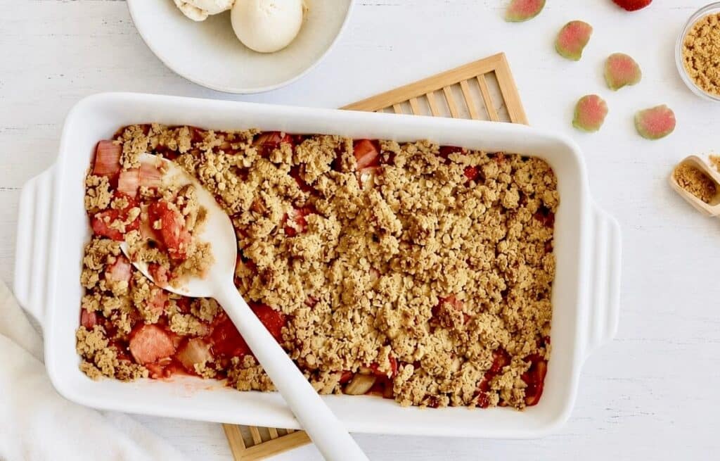 Veganer Rhabarber-Erdbeer-Crumble in einer Auflaufform auf einem Holztisch mit einer Schüssel mit 3 Kugeln Vanilleeis.