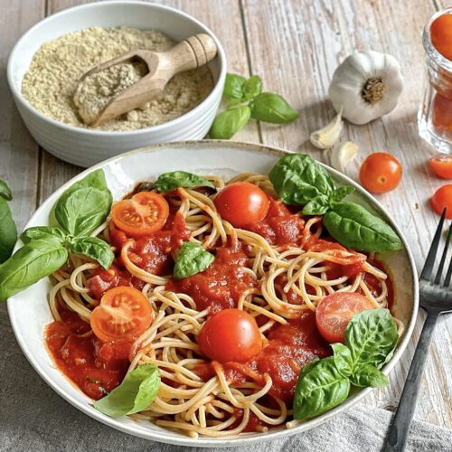 Vegane Tomaten Basilikum Pasta auf einem Teller mit Cashew-Parmesan in einer Schüssel.