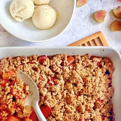 Veganer Rhabarber-Erdbeer-Crumble in einer Auflaufform auf einem Holztisch mit einer Schüssel mit 3 Kugeln Vanilleeis.