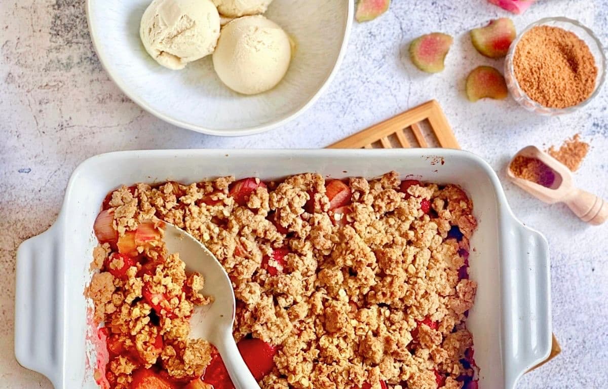 Veganer Rhabarber-Erdbeer-Crumble in einer Auflaufform auf einem Holztisch mit einer Schüssel mit 3 Kugeln Vanilleeis.