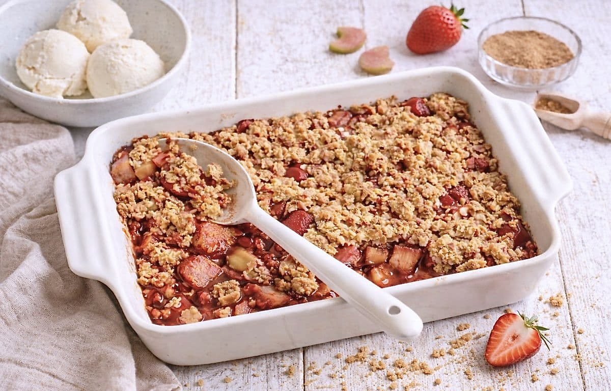 Veganer Rhabarber-Erdbeer-Crumble in einer Auflaufform auf einem Holztisch mit einer Schüssel mit 3 Kugeln Vanilleeis.