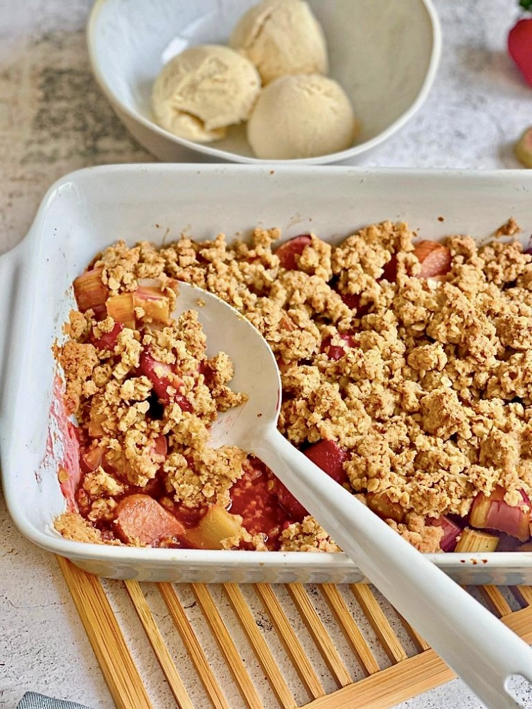 Veganer Rhabarber-Erdbeer-Crumble in einer Auflaufform auf einem Holztisch mit einer Schüssel mit 3 Kugeln Vanilleeis.