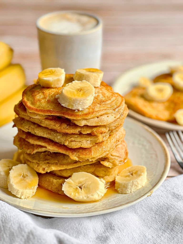 Vegane Bananen-Pancakes mit Ahornsirup, der von oben übergegossen wird, gestapelt auf einem Teller.