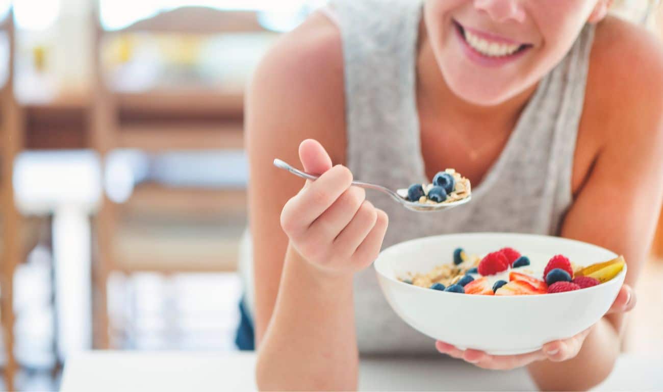 Eine lächelnde Frau, die eine Schale Müsli mit Obst isst und somit eine gesunde vegane Ernährung umsetzt.