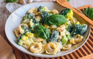 Vegane Tortellini mit Käsesoße, Brokkoli und Basilikum auf einem Teller auf einem Holzbrett und mit einer Holzgabel.