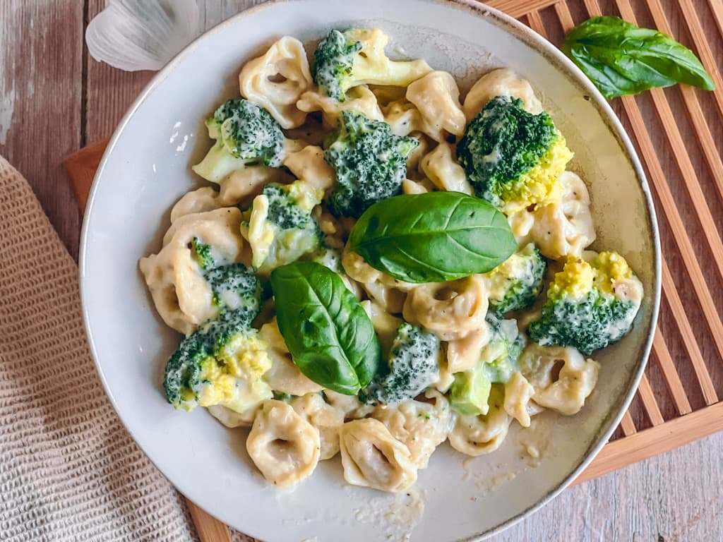 Vegane Tortellini mit Brokkoli in Käsesoße auf einem Teller auf einem Holzbrett.