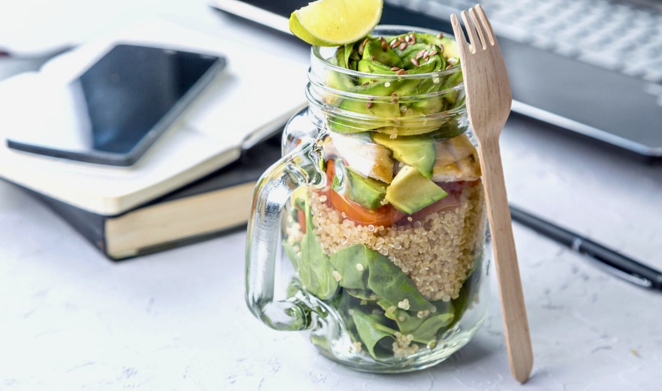 Veganer Salat in einem Glas mit einer Holzgabel vor Büchern, einem Smartphone und einer Tastatur.