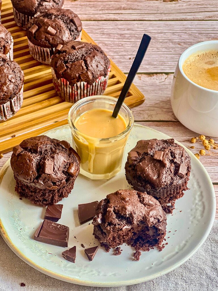 Vegane Schokomuffins (Double Chocolate Muffins) auf einem Teller mit Mandelmus und auf einem Holzbrett, daneben eine Tasse Kaffee.