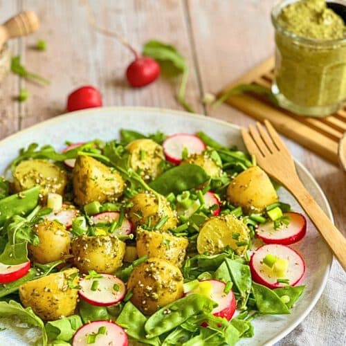 Veganer Kartoffelsalat mit Pesto, Rucola, Radieschen, Zuckerschoten und Frühlingszwiebeln auf einem Teller.