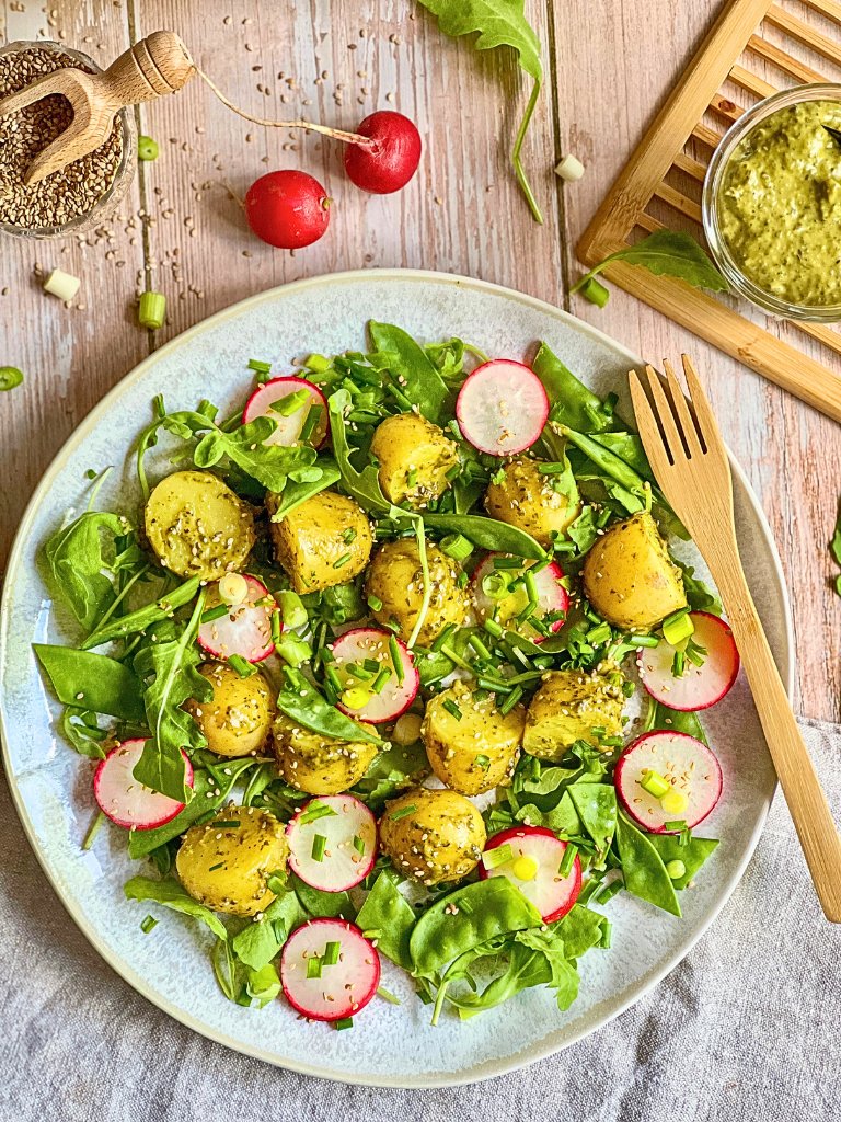 Veganer Kartoffelsalat mit Pesto, Rucola, Radieschen, Zuckerschoten und Frühlingszwiebeln auf einem Teller.