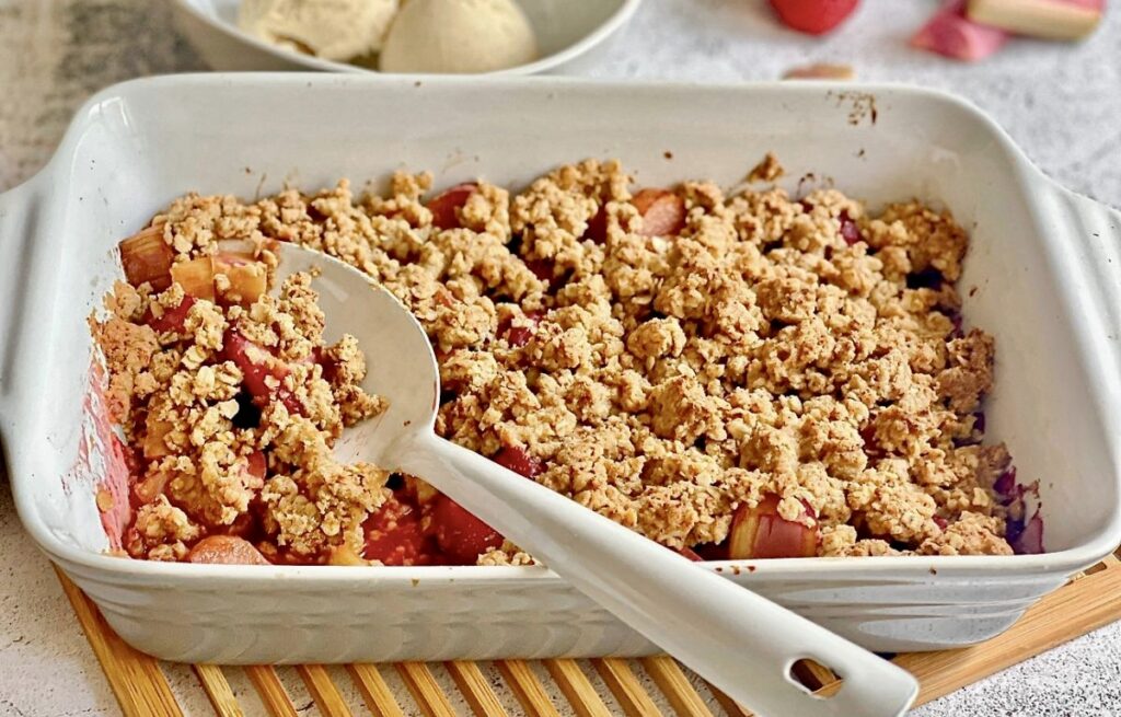 Veganer Rhabarber-Erdbeer-Crumble in einer Auflaufform auf einem Holztisch mit einer Schüssel mit 3 Kugeln Vanilleeis.