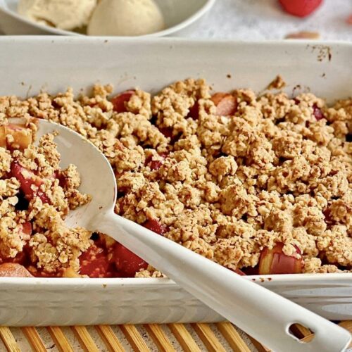 Veganer Rhabarber-Erdbeer-Crumble in einer Auflaufform auf einem Holztisch mit einer Schüssel mit 3 Kugeln Vanilleeis.