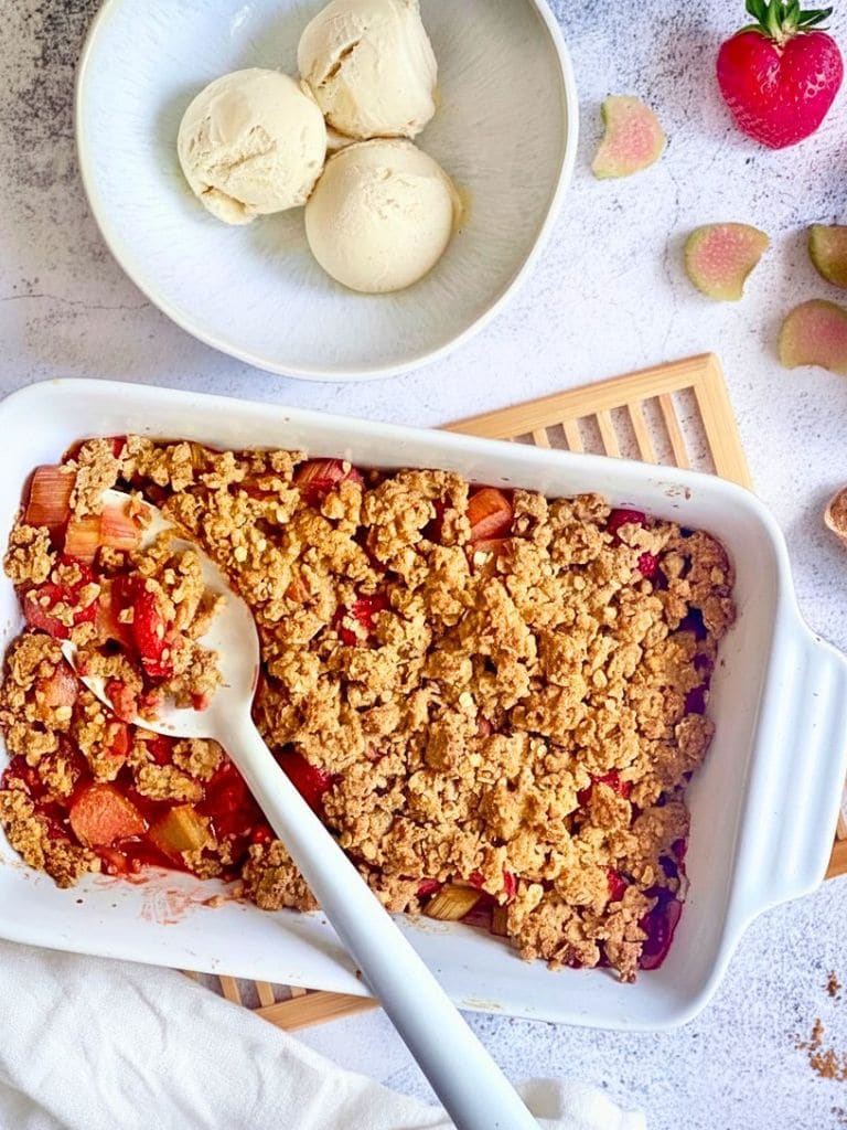 Veganer Rhabarber-Erdbeer-Crumble in einer Auflaufform auf einem Holztisch mit einer Schüssel mit 3 Kugeln Vanilleeis.