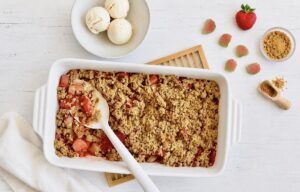 Veganer Rhabarber-Erdbeer-Crumble in einer Auflaufform auf einem Holztisch mit einer Schüssel mit 3 Kugeln Vanilleeis.