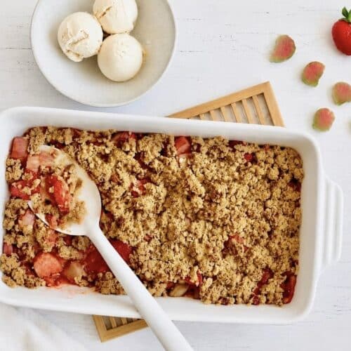 Veganer Rhabarber-Erdbeer-Crumble in einer Auflaufform auf einem Holztisch mit einer Schüssel mit 3 Kugeln Vanilleeis.