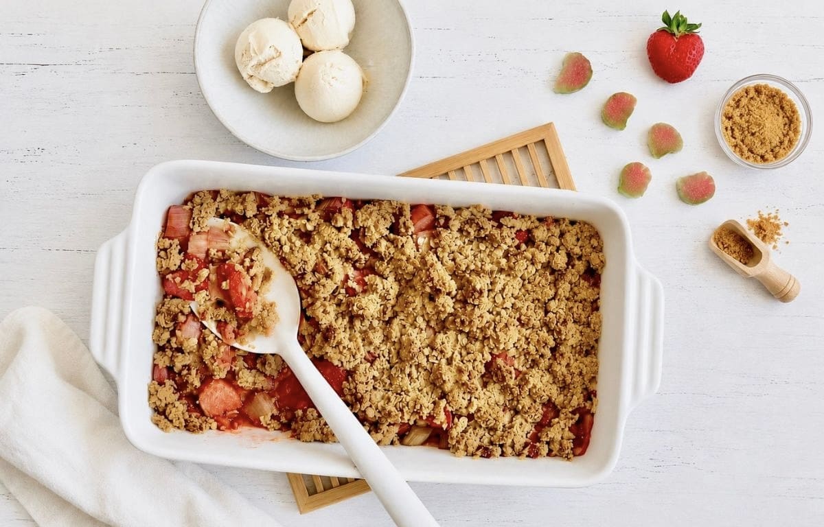 Veganer Rhabarber-Erdbeer-Crumble in einer Auflaufform auf einem Holztisch mit einer Schüssel mit 3 Kugeln Vanilleeis.