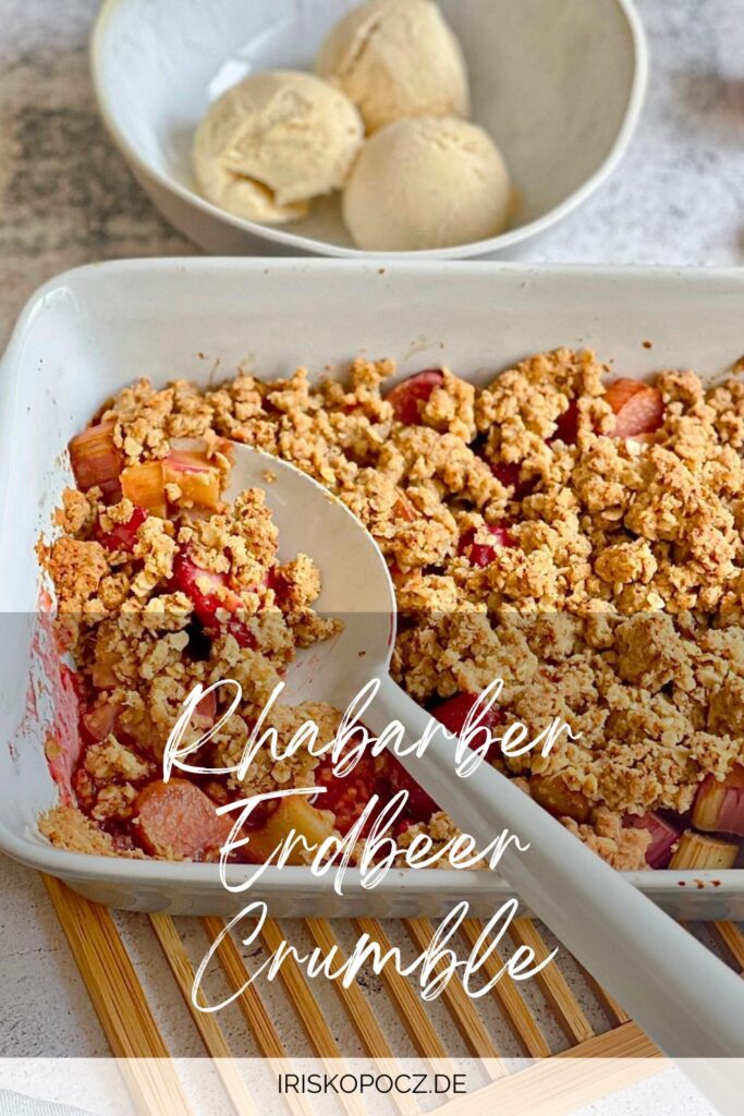 Veganer Rhabarber-Erdbeer-Crumble in einer Auflaufform auf einem Holztisch mit einer Schüssel mit 3 Kugeln Vanilleeis-Pin.